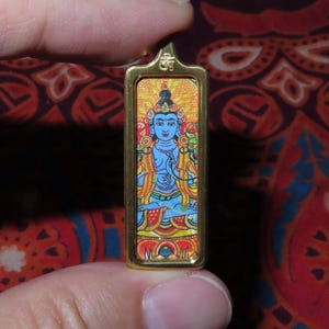 Puede incluir: Colgante rectangular dorado con una colorida ilustración de una deidad de piel azul, con corona y vestimenta ornamentada. El colgante, con un pequeño lazo en la parte superior, está sujeto entre los dedos.