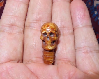May include: A small, detailed skull carving in shades of brown and tan. The skull has visible eye sockets, a nasal cavity, and a detailed jawline. The carving is held in an open hand, showcasing its size and intricate design.