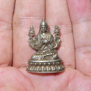 May include: A small, silver-coloured statue of a seated figure wearing a pointed hat and ornate clothing. The figure is holding its hands in prayer and is flanked by two decorative floral elements. The statue sits on a detailed, round base.