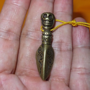 May include: A brass-toned metal pendant featuring a carved skull head with a wide, toothy grin at the top, transitioning into a tapered, teardrop-shaped body decorated with etched swirling patterns, attached to a thin, bright yellow cord.