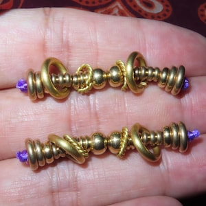 May include: Two gold-coloured beaded jewellery components with purple thread ends. Each component features a series of rounded beads, rings, and twisted wire accents. The components are displayed against a neutral background.