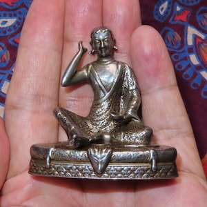 May include: A small, silver-colored statue of a seated figure with one hand raised. The figure is on a detailed base with a decorative border. The statue is held in a person's hand, with a patterned fabric in the background.