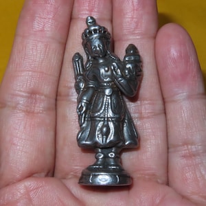 Op de afbeelding: Een klein, metaalachtig zilverkleurig beeldje dat een staande godheid of koninklijk figuur voorstelt, gekleed in een versierde kroon en traditionele gewaden. Het figuur houdt een cilindervormig object in één hand en staat op een ronde voet met gedetailleerde reliëfpatronen.