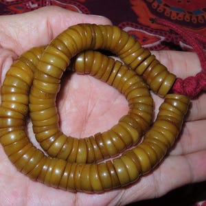 May include: A strand of prayer beads made of light brown, cylindrical beads. The beads are strung together and tied with a red cord. The beads are held in a hand.