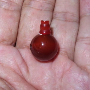 Puede incluir: Una pequeña cuenta redonda, de color rojo oscuro, con una parte superior cilíndrica. La cuenta tiene un acabado brillante y parece estar hecha de un material liso y pulido. La cuenta se sostiene en una mano.