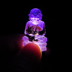 May include: A small, purple, glass figurine of a seated Buddha. The figurine is illuminated from below, casting a purple glow.