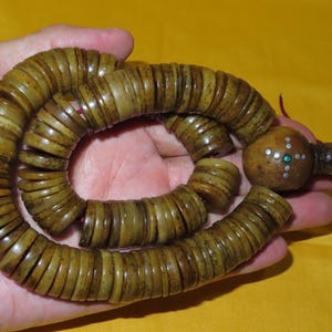 May include: A string of flat, circular, brownish-tan beads arranged in a spiral pattern. The beads are threaded on a red cord, ending in a larger, rounded, textured bead featuring a small cross inlay with silver-colored dots and a green center.