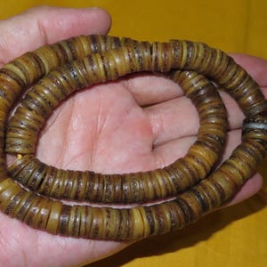 May include: A circular strand of small, flat, disc-shaped beads in shades of tan, brown, and dark amber. The beads are strung together on a thin yellow cord, with a single metallic silver-toned spacer bead near the end of the loop.