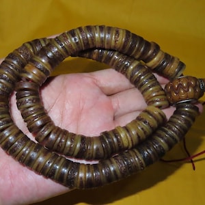 May include: A circular strand of brown, disc-shaped beads with textured, dark brown patterns. The beads are strung together, ending in a carved, oval-shaped brown bead with a silver-toned metal accent and a thin red cord attached to the end.
