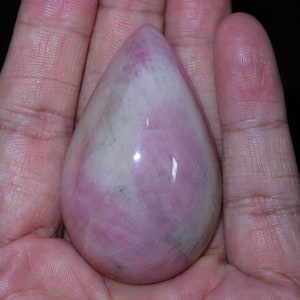 Puede incluir: Un colgante de piedra preciosa en forma de lágrima de color rosa y blanco. El colgante es liso y pulido, con un tono rosa claro y vetas blancas.