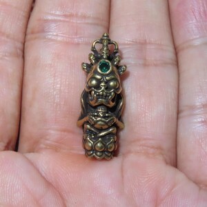 Könnte beinhalten: Ein detaillierter, bronzefarbener Anhänger mit einer stilisierten Gottheit mit grimmigem Ausdruck und einem grünen Edelstein. Der Anhänger weist komplizierte Details auf, darunter eine Krone und eine Figur, die eine Lotusblume hält. Der Anhänger wird in einer Hand gehalten.