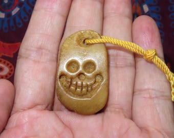 May include: A carved, tan-colored pendant with a smiling skull design. The pendant is rectangular with rounded edges and a hole at the top. A yellow cord is attached. The pendant is held in a person's hand.