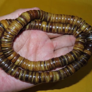 May include: A strand of circular, disc-shaped beads in varying shades of brown, tan, and dark amber. The beads are strung together on a thin cord, creating a continuous loop with a textured, weathered appearance and natural color variations.