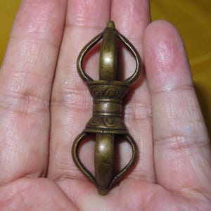 May include: A small, antique-style bronze vajra ritual object featuring a symmetrical design with curved prongs extending from a central, engraved cylindrical handle. The metal has a darkened, weathered patina with visible texture and detailing.