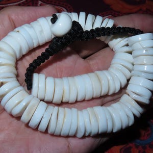 Op de afbeelding: Een witte schelpenketting met een zwart koord en een witte kraal. De ketting is gemaakt van veel kleine, ronde schelpen.