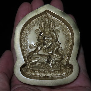 May include: A gold-coloured metal mould for casting a deity statue. The mould is shaped like a teardrop and features a detailed design of a deity with a crown and a fierce expression. The deity is holding a weapon in one hand and a bell in the other. The mould is surrounded by a decorative border.