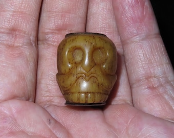 May include: A small, carved skull bead made of brown bone with a metal ring around the top. The bead is detailed with carved eyes, nose, and mouth.