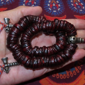 Può includere: Una collana di perline di preghiera mala rosso scuro con una serie di piccole perline rotonde. La collana è tenuta in una mano, con accenti metallici color argento e corda rossa e blu.