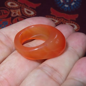 May include: A smooth, orange ring with a rounded design. The ring is a solid band with a circular opening in the centre. The colour is a warm, vibrant orange, and the material appears to be polished stone or resin.
