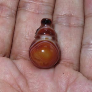 May include: A small, brown and orange agate pendant with a rounded bottom and a bulbous shape. The pendant has a dark brown top and lighter brown and white banding throughout. The agate is held in a person's hand.
