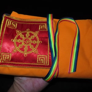 Puede incluir: Un objeto de tela naranja con una sección bordada en rojo con un diseño de rueda dorada. Se adjunta una correa a rayas arcoíris. El artículo está doblado y parece ser un textil religioso o ceremonial.