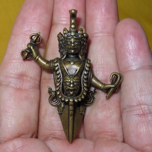 Puede incluir: Un colgante de metal en tono bronce que representa una deidad de múltiples rostros con una expresión feroz. La figura sostiene un vajra en una mano y una serpiente en la otra, con un rostro más pequeño en el pecho, terminando en una punta de daga.