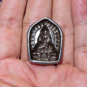 May include: A silver pendant showing a seated figure in an arched frame. The figure is in a meditative pose, with detailed clothing and surroundings. The pendant is held in an open hand. The pendant is likely made of silver.