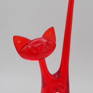 May include: A transparent red cat-shaped decorative object. The cat has large ears, a small face, and a long, curved tail. The object is made of a glossy material and stands upright.