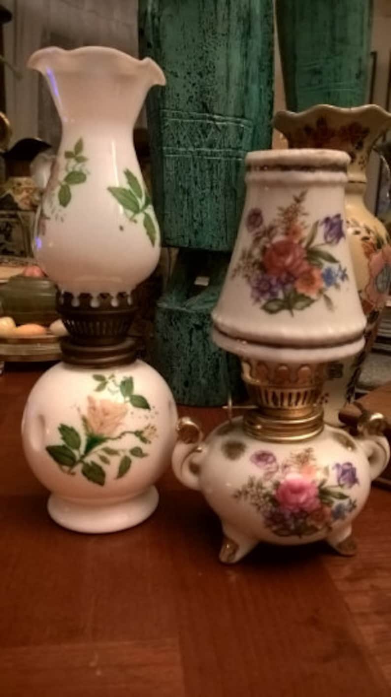 May include: Two vintage porcelain oil lamps with floral designs. The lamps are white with gold accents and feature delicate floral patterns in shades of pink, yellow, and green. The lamps are on a wooden surface.