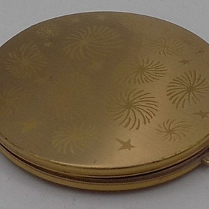 Puede incluir: Un espejo compacto dorado con un diseño de fuegos artificiales y estrellas. El espejo está cerrado.