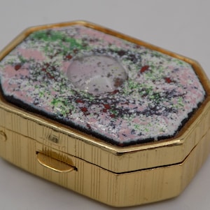 Könnte beinhalten: Eine goldene achteckige Kompaktdose mit einem Scharnierdeckel. Der Deckel ist mit einem gesprenkelten Muster in Rosa, Grün und Weiß verziert, mit einem klaren runden Zentrum.
