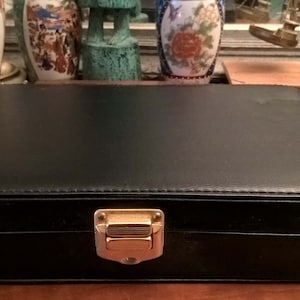 May include: A rectangular black jewelry box with a gold-colored clasp. The box is made of a smooth, possibly leather-like material. The box is closed and sits on a wooden surface.