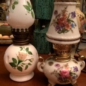 May include: Two vintage oil lamps with floral designs. The lamp on the left is white with a pink rose and green leaves. The lamp on the right is white with a pink, yellow, and purple floral design.