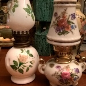 May include: Two vintage oil lamps with floral designs. The lamp on the left has a white base with a single rose and green leaves. The lamp on the right has a white base with a floral pattern in shades of pink, yellow, and blue.