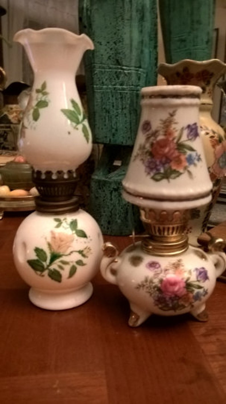 May include: Two vintage oil lamps with floral designs. The lamp on the left has a white base with a single rose and green leaves. The lamp on the right has a white base with a floral pattern in shades of pink, yellow, and blue.