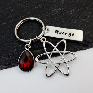 May include: Silver keyring with a red teardrop-shaped crystal, an atom charm, and a personalised tag that reads "George".