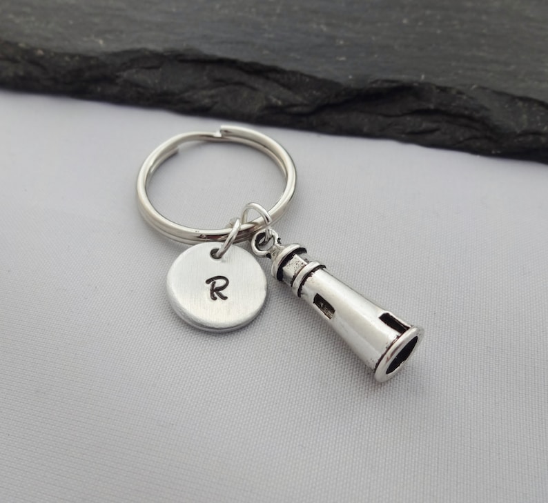 Puede incluir: Llavero de plata con un dije de faro y un dije redondo con la letra R grabada.