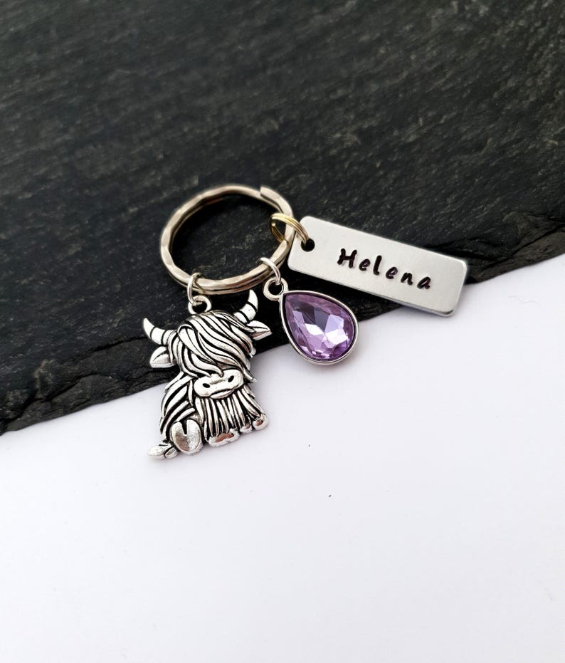 Puede incluir: Un llavero plateado con un dije de vaca de las Tierras Altas, una gema morada en forma de l&aacute;grima y una etiqueta rectangular con el nombre "Helena" grabado. El llavero est&aacute; sujeto a un anillo plateado.
