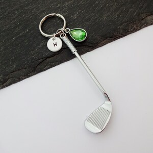 Puede incluir: Llavero de club de golf plateado con un dije de cristal verde y un dije plateado con la letra H.