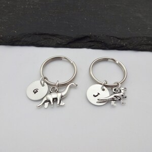 May include: Two silver keychains with dinosaur charms. One keychain has a brontosaurus charm and a round tag with the letter "G" on it. The other keychain has a T-Rex charm and a round tag with the letter "J" on it.