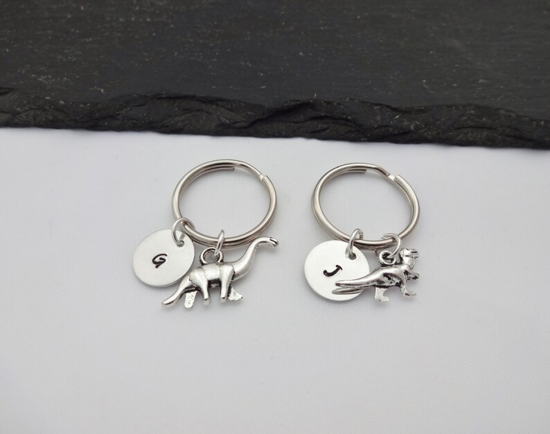 May include: Two silver keychains with dinosaur charms. One keychain has a brontosaurus charm and a round tag with the letter "G" on it. The other keychain has a T-Rex charm and a round tag with the letter "J" on it.