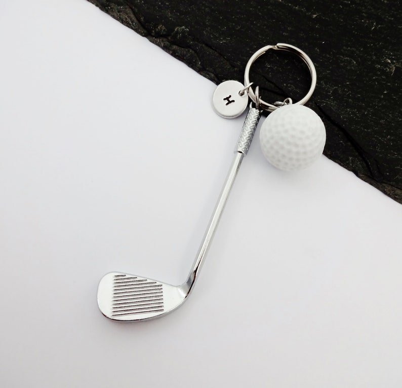 Puede incluir: Llavero de club de golf plateado con una bola de golf blanca y un dije plateado con la letra H.