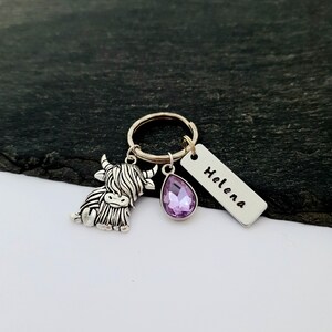 Puede incluir: Un llavero plateado con un dije de vaca de las Tierras Altas, una gema morada en forma de l&aacute;grima y una etiqueta rectangular con el nombre "Helena" grabado. Ideal para llaves o como accesorio decorativo.