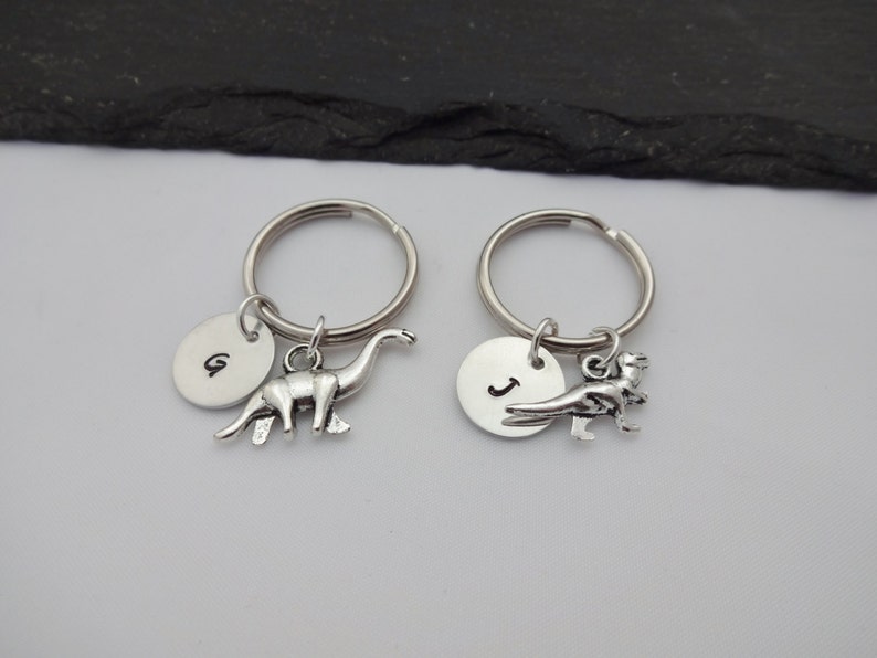 May include: Two silver keychains with a dinosaur charm and a round disc with an initial. The keychain on the left has a brontosaurus charm and the disc has the initial "G". The keychain on the right has a T-Rex charm and the disc has the initial "J".