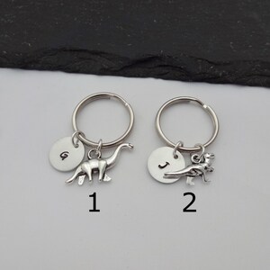 May include: Two silver keychains with dinosaur charms. The first keychain has a letter G charm and a brachiosaurus charm. The second keychain has a letter J charm and a T-Rex charm.