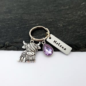 Puede incluir: Un llavero plateado con un dije de vaca de las Tierras Altas, una gema morada en forma de l&aacute;grima y una etiqueta rectangular grabada con el nombre "Helena". El llavero est&aacute; en un anillo plateado.