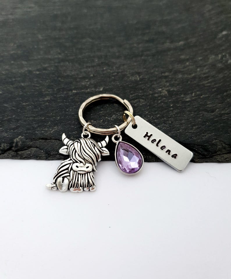 Puede incluir: Un llavero plateado con un dije de vaca de las Tierras Altas, una gema morada en forma de l&aacute;grima y una etiqueta rectangular grabada con el nombre "Helena". El llavero est&aacute; en un anillo plateado.