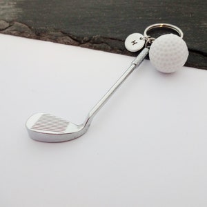 Puede incluir: Un llavero de palo de golf plateado con un abalorio de bola de golf blanca y un abalorio plateado con la letra "H" en &eacute;l.