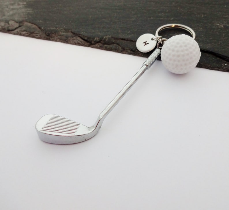 Puede incluir: Un llavero de palo de golf plateado con un abalorio de bola de golf blanca y un abalorio plateado con la letra "H" en &eacute;l.
