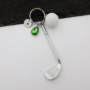 Puede incluir: Un llavero de palo de golf plateado con una bola de golf blanca, un amuleto de cristal verde y un amuleto plateado con la letra "H" en &eacute;l.